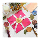 Pink envelope with a gold wax seal surrounded by various seals and a candle on a textured surface.