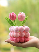 Hand holding a pink crochet berry and tulip toy against a blurred green background