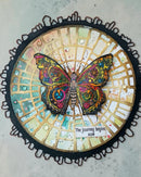 Decorative circular piece with a colorful butterfly and 'The Journey Begins Now' text on a textured surface.
