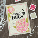 Card with 'Sending Hugs' text and pink roses on a wooden surface with stamps and stencils.
