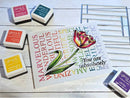 Card with a tulip illustration and赞美 text, surrounded by Gina K. Designs stamps on a wooden surface.