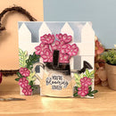 Card with pink flowers, a watering can, and 'You're Blooming Lovely' text on a wooden surface.