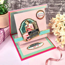Greeting card with decorative elements and text on a pink surface with flowers and a gift box.