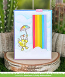 Card with a duck holding an umbrella and rainbow, placed on a wooden block with plants in the background.