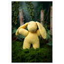 Yellow crochet bunny toy on a green grass background with plants and branches.