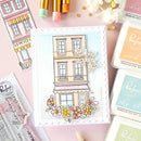 Card with a building illustration and flowers, surrounded by stamping supplies on a pink background
