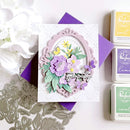 Handmade card with floral design and purple accents, surrounded by floral elements and craft supplies.
