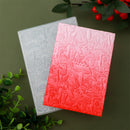 Two embossed cardstocks, one gray and one pink, on a green background with foliage.