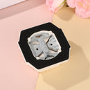 Metallic butterfly-shaped earrings in a black hexagonal box on a pink and beige background with pink flowers.