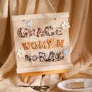 Decorative sign with floral letters spelling 'GRACE WOMEN MORAL' on a wooden stand against a beige curtain.