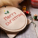 Embroidery hoop with 'Merry Christmas' text and Christmas tree design on a wooden surface with spools of thread.