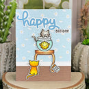 Birthday card with cartoon cats and a fishbowl design, placed on a wooden surface with plants in the background.