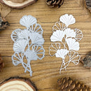 Two white leaf-shaped metal cutting dies on a wooden surface with pine cones.
