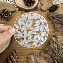Decorative metal cutting die with leaf pattern held over a wooden surface with pinecones and branches.