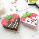 Two strawberry-shaped chocolate boxes on a white surface with colorful candies around.