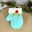 Blue mitten ornament with holly and red button on a wooden surface with pine branches.