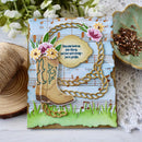 Decorative card with cowboy boot, flowers, and music notes on a white surface with floral and yarn background.