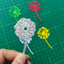 Metal dandelion flower cutout held by a hand on a green cutting mat with colorful paper dandelions.