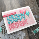 Birthday card with balloons and text on a wooden surface with crafting tools.
