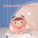Hand knitted doll with pink outfit and brown hair on a decorative stand with 'HAND KNITTED DOLL' text above.