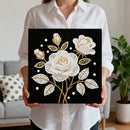 Person holding a decorative painting with white and gold floral design on a black background.