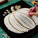 Hand applying pearls to a decorative pumpkin design on a black surface with tools around.