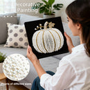Woman holding a completed pearl painting of a pumpkin on a couch with decorative pillows.