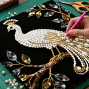 Handmade peacock design with pearls and gold accents on a black background, with a hand using a pink tool.