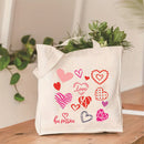 White tote bag with heart designs and 'be mine' text on a wooden surface with plants in the background