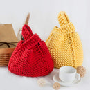 Two chunky knit drawstring handbags, one in bright red and one in soft yellow, styled on a white surface with a white teacup, saucer, small woven balls, and a brown paper bag in the background.