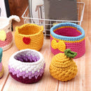 A set of handmade crocheted storage baskets and containers in various colorful patterns, including yellow with a red heart, pink and blue with a yellow bow, purple and white, and yellow with a green leaf, arranged on a wooden surface.