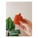 Hand holding a small orange knitted item against a neutral background with a plant in the corner.