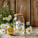 Glass jar with wooden lid and straw, filled with water and lemon slices, on a wooden surface with flowers in the background.