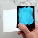 Hand holding a small square of paper with blue paint swatch against a marble background