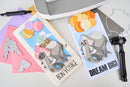 Two greeting cards with elephant designs and text, surrounded by crafting tools on a light surface.