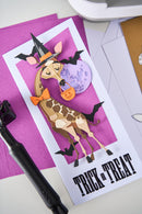 Halloween-themed greeting card with a giraffe wearing a witch hat and holding a pumpkin, on a pink and white striped background.