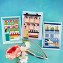 Three colorful greeting cards with building designs on a blue background, accompanied by a pair of scissors and flowers.