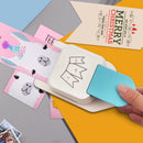 Hand using a white card puncher on colorful cards with a 'Merry Christmas' card in the background.