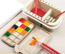 Artistic set with watercolor paint tray, brush, and container on a white surface.