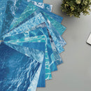 Stack of blue water-patterned paper sheets on a gray surface with a plant in the corner.
