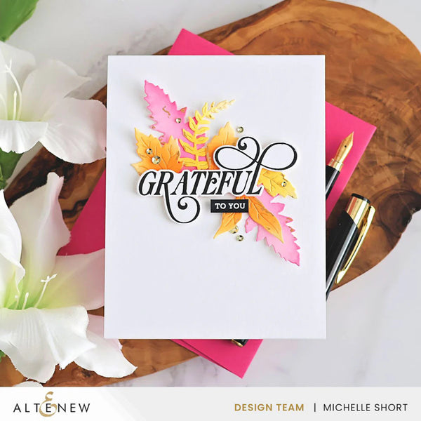 Greeting card with 'GRATEFUL TO YOU' text and autumn leaves on a wooden surface with flowers and stationery.