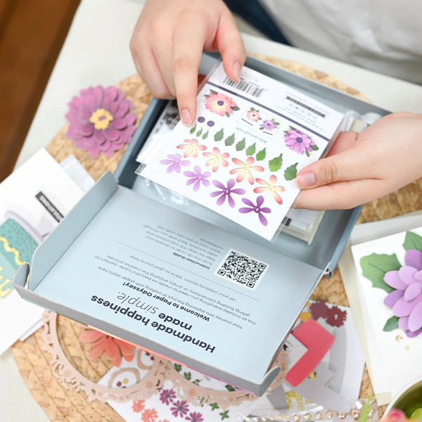 Hands opening a box of floral stickers with a QR code on a table.