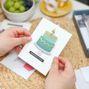 Hand holding a card with a cake illustration and 'Congratulations' text on a woven surface.