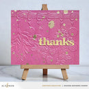 Pink card with gold embossed design and 'thanks' text on a wooden easel.