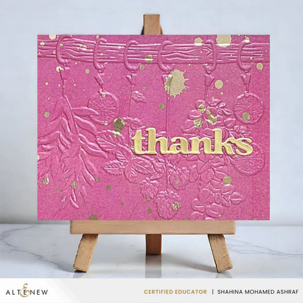 Pink card with gold embossed design and 'thanks' text on a wooden easel.