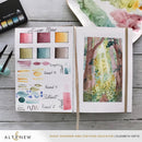 Open art supply book with watercolor samples and a forest scene on a wooden surface, featuring Altenew branding.