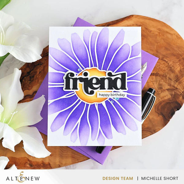 Greeting card with purple flower design and 'friend happy birthday' text on a wooden surface with flowers and a pen.