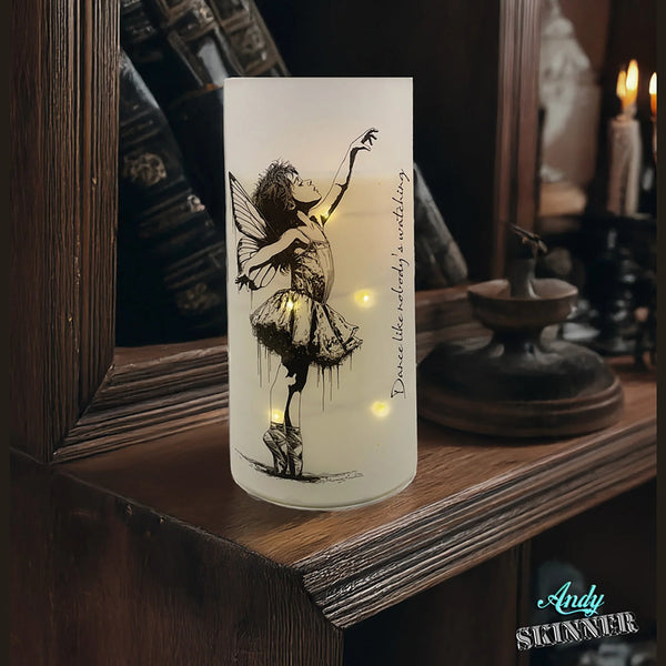 Decorative candle with fairy design and string lights on a wooden surface.