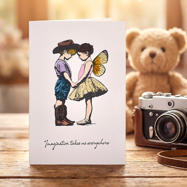 Greeting card with illustration of a boy and girl holding hands, surrounded by a teddy bear and camera on a wooden surface.