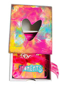 Colorful gift box with a heart-shaped window and 'Moments' text, featuring a pink ribbon.
